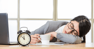 woman falling asleep on a desk next to an alarm clock because she haven't slept since the time change
