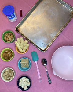 spread of ingredients for super easy valentine's dessert board. baking sheet, bowl, spatula, spoon, frosting, food coloring, cookies and treats.